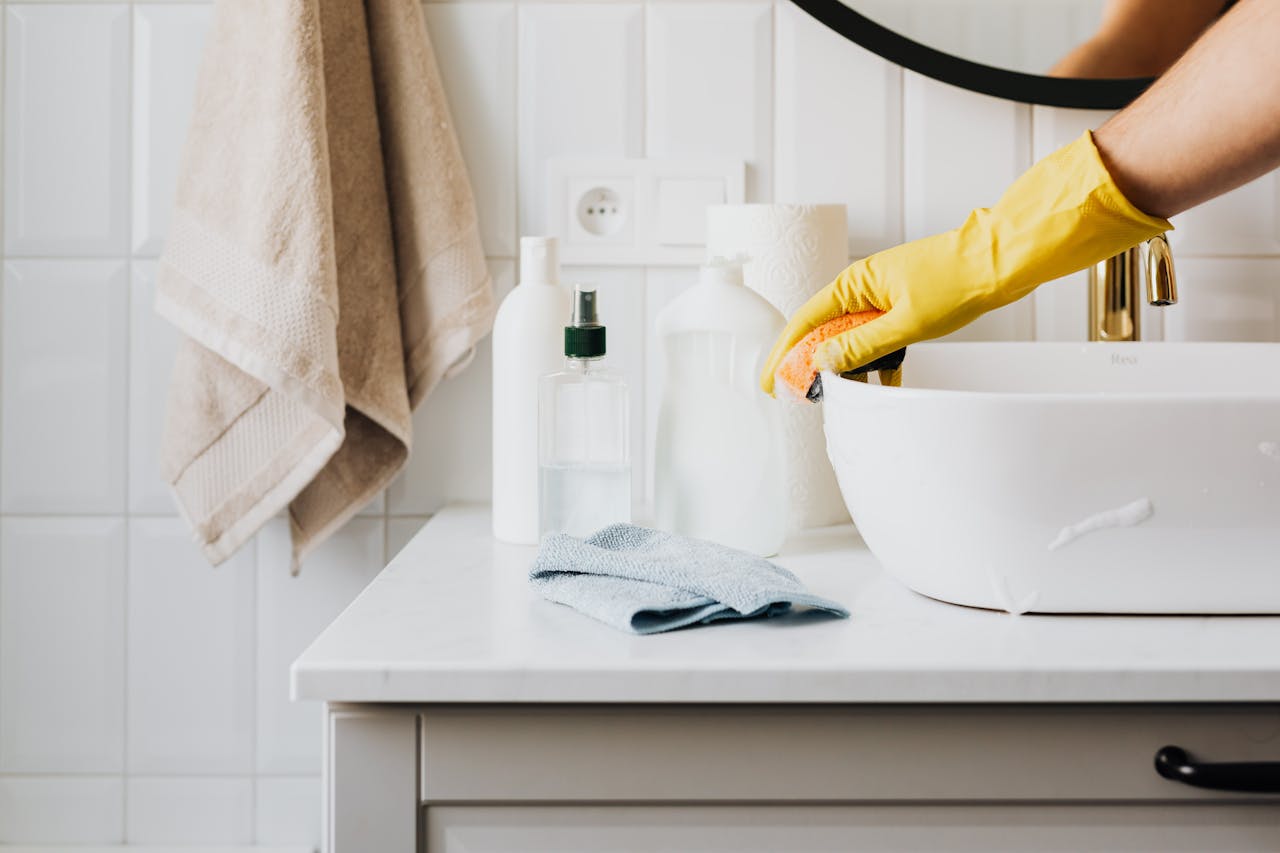 Person cleaning a bathroom sink with yellow gloves and cleaning products on a countertop.